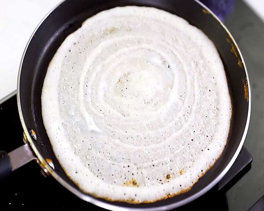 Spreading batter in a circular motion to cook crispy dosa on a pan.