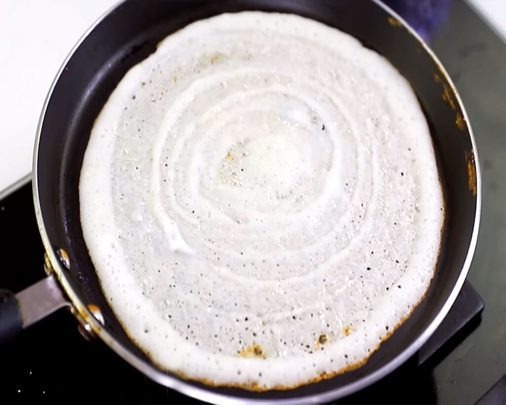 Dosa cooking on the tawa with golden brown edges lifting, indicating it’s ready to be flipped and served.
