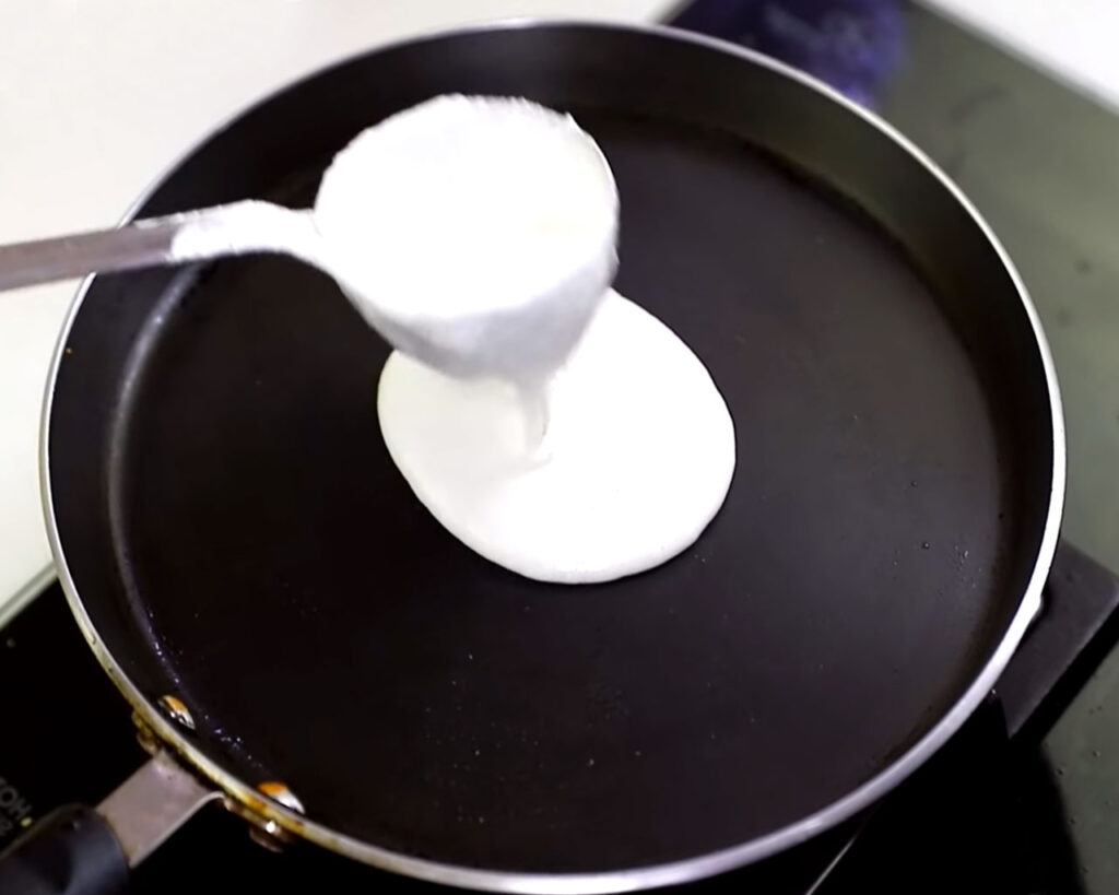Dosa batter being poured in the center of a hot non-stick tawa, preparing to be spread into a thin layer.