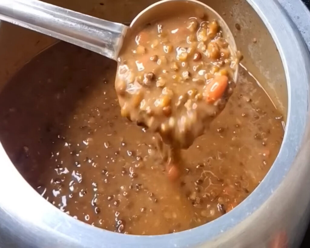 Soft cooked lentils being mashed with a wooden masher.