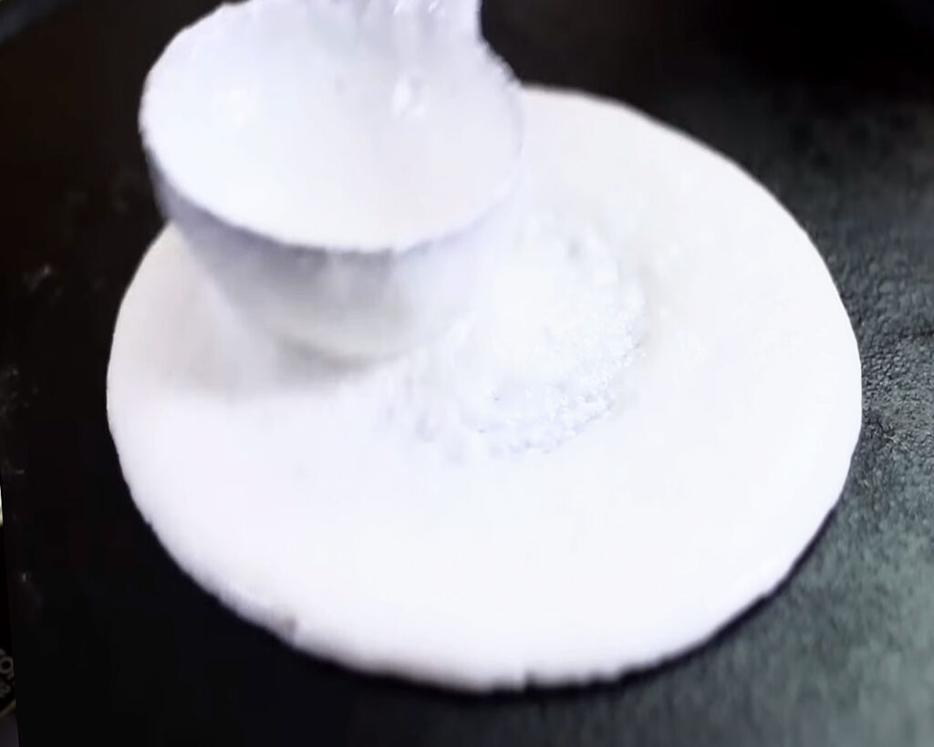 Using the back of a ladle to spread dosa batter in a circular motion, forming a thin, even layer on the tawa.