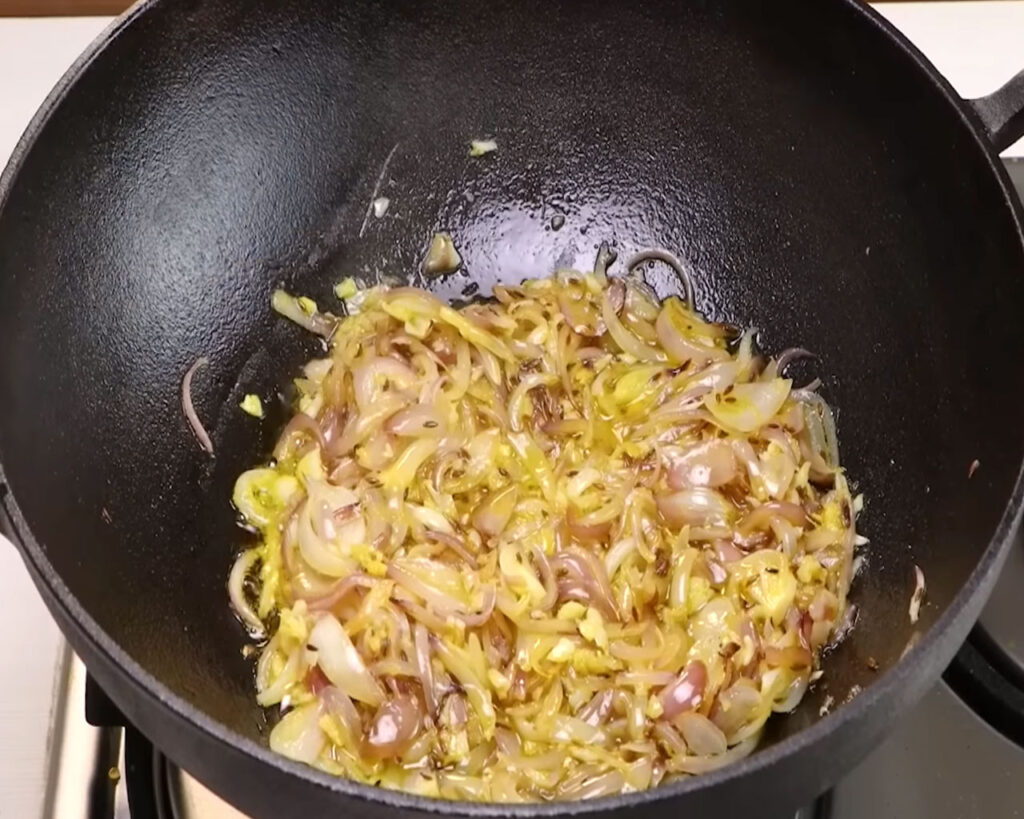 Adding ginger garlic paste to golden fried onions in a kadhai for rich aroma in rajma curry.