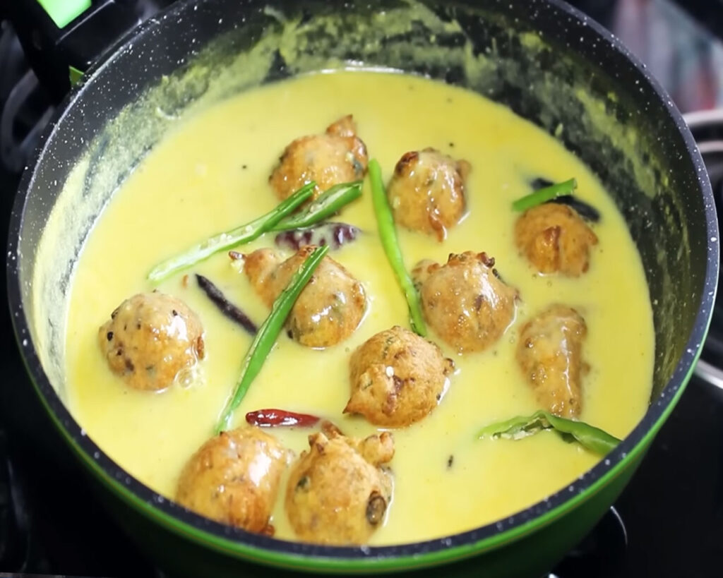 Adding fried pakoras into the kadhi to soak up flavors and simmer.