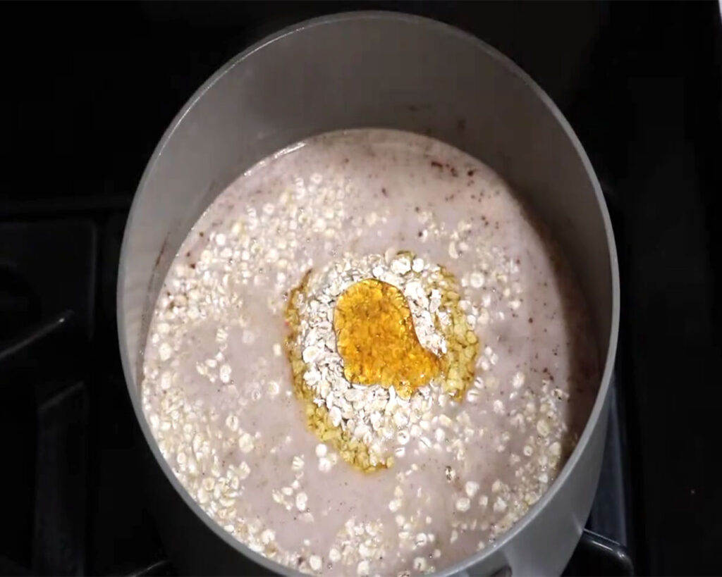 One cup of oatmeal being stirred into coconut milk in a pot, with salt added for flavor and honey drizzled on top to sweeten the coconut milk oatmeal mixture.