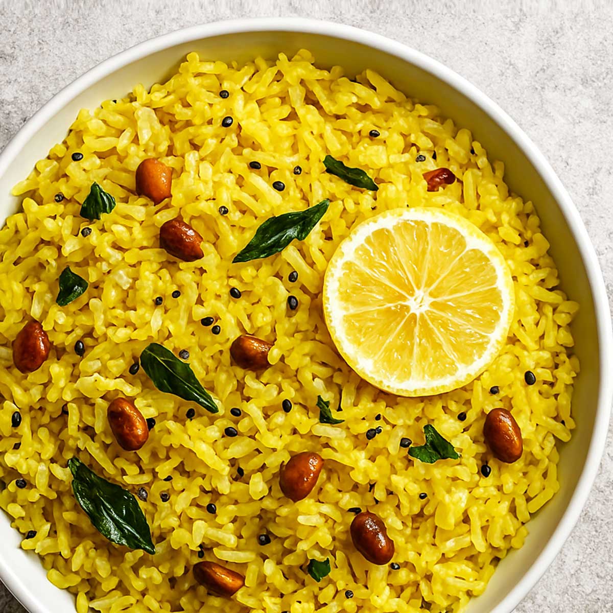 Bright yellow lemon rice on a white plate, flavored with turmeric, curry leaves, green chilies, and peanuts, served with a fresh lemon slice.