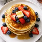 Soft homemade eggless pancakes stacked and drizzled with honey, topped with sliced strawberries
